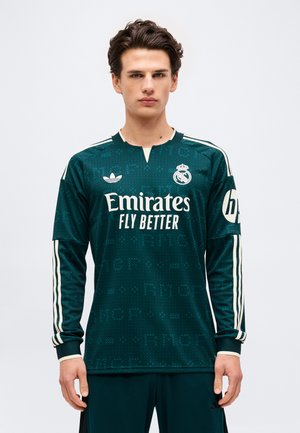 Giovane uomo che indossa una maglia da calcio a maniche lunghe del Real Madrid verde scuro con strisce bianche e il logo dello sponsor "Emirates Fly Better".