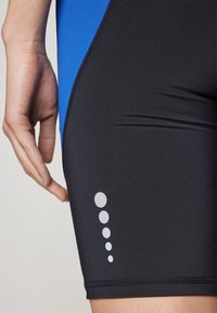 Pantaloni da bagno neri con accenti blu, dotati di un pannello laterale testurizzato e un motivo circolare bianco lungo la coscia sinistra. Tessuto liscio.