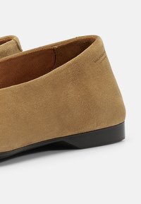 Sømsko i mokka i tan, med rund tå, lav sort hæl og tekstureret inderside. Minimal branding på bagsiden.