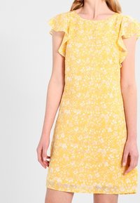 Kvinna i en gul blommig klänning med korta volangärmar, stående mot en enkel vit bakgrund.