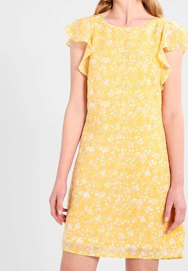 Kvinna i en gul blommig klänning med korta volangärmar, stående mot en enkel vit bakgrund.