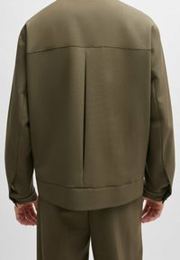 Olijfgroene jas met een gestructureerde afwerking, voorzien van een plooi aan de achterkant, brede manchetten en een rechte zoom. Strak, minimalistisch ontwerp.