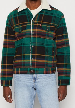 Mann trägt eine grün-schwarz-braun karierte, sherpa-gefütterte Jacke über einem weißen Hemd, kombiniert mit hellblauen Jeans.