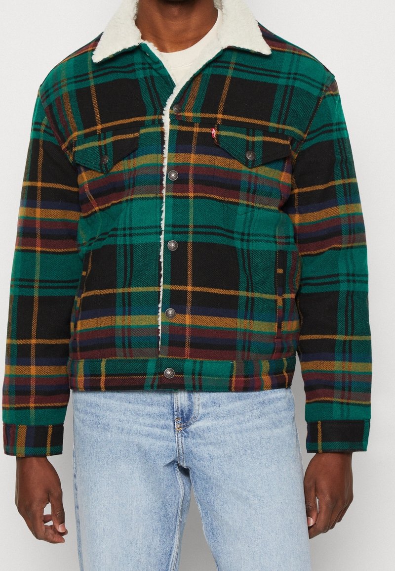 Homme portant une veste à carreaux verte, noire et marron doublée en sherpa, par-dessus une chemise blanche, associée à un jean bleu clair.