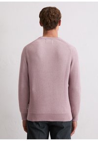 Man facing away wearing a light purple textured knit sweater and dark pants against a plain white wall background.