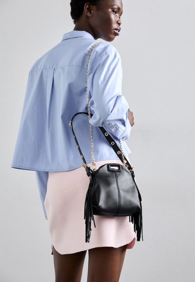 Sac en cuir noir avec des détails en frange, une chaîne en or et un design structuré. Le modèle porte une chemise bleu clair et une jupe rose.