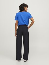 Blauwe katoenen T-shirt met een ronde hals, gecombineerd met hoge zwarte wijde broeken. Het model heeft krullend haar en wordt van achteren getoond.