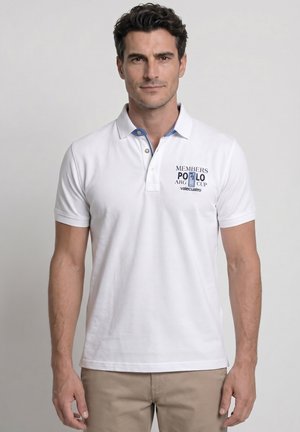 Hombre con una camiseta polo blanca con el texto "Members Polo Arg Cup", de pie frente a un fondo gris claro liso.