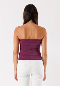 Top viola senza spalline con una finitura testurizzata e lucente; design aderente. Indossato con jeans bianchi con tasche posteriori.