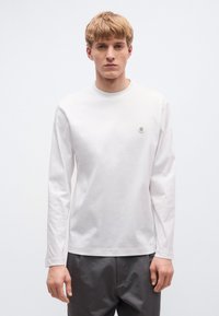 Hosszú ujjú fehér póló puha pamutból, kerek nyakkivágással és egy apró emblémával a mellkason, sötétszürke nadrággal párosítva.