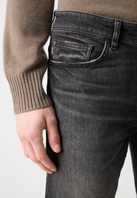 Main gauche à côté d'un jean gris foncé avec une petite étiquette "BOSS" visible, associée à un pull beige à manches côtelées.