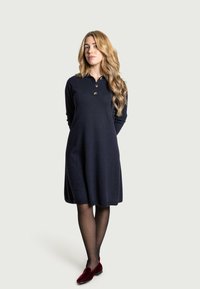 Donna con lunghi capelli biondi mossi, che indossa un vestito blu navy, collant neri e mocassini di velluto rosso, in piedi con le mani dietro la schiena.