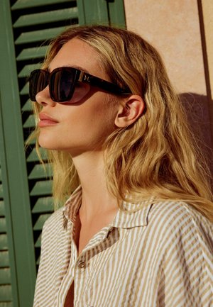 Blondýna s tmavými velkými slunečními brýlemi a béžovým pruhovaným tričkem stojící u zelených okenic za teplého slunečního svitu.