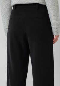 Pantalon noir à la texture lisse, avec deux poches arrière et une coupe ajustée. Porté avec un pull texturé gris clair.