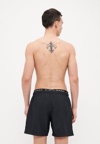 Calções de natação pretos com uma cintura elástica, apresentando uma faixa com o logo. O modelo é mostrado de costas, exibindo uma tatuagem floral na parte superior das costas.