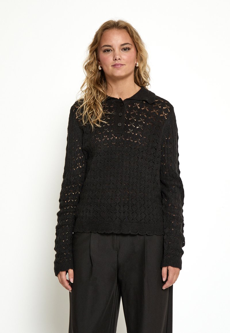 Zwarte gebreide pullover met een knoopsluiting bij de kraag, gekenmerkt door een gestructureerd opengewerkte patroon en een gekartelde zoom, gecombineerd met high-waisted broeken.