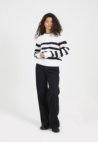 Biały sweter robiony na drutach z czarnymi poziomymi paskami, w połączeniu z czarnymi spodniami o szerokich nogawkach. Sweter ma teksturowany wzór i ściągacze na mankietach.