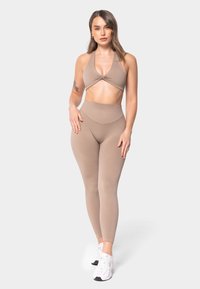 Sujetador deportivo beige con detalle de nudo, combinado con leggings beige de cintura alta. La modelo lleva zapatillas blancas; tejido suave, diseño ajustado.
