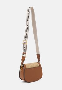 Ruskea olkalaukku, jossa on kudottu läppä, kultaiset yksityiskohdat ja säädettävä beige olkahihna, jossa on mustalla tekstillä "KARL LAGERFELD".