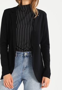 Blazer negro de tejido suave con frente abierto, combinado con un cuello alto de rayas negras y blancas. Vaqueros de mezclilla claros de tiro alto.