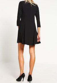 Robe noire à manches longues, jupe évasée et corsage ajusté. Confectionnée dans un matériau lisse avec un design épuré et minimaliste. Associée à des talons noirs.