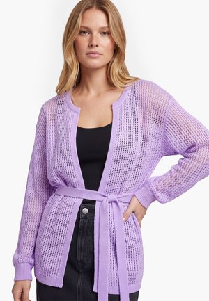 Vrouw draagt een lichtpaars opengebreid vest met een riem gestrikt in de taille over een zwarte top en donkere hoogtaillepantalon.