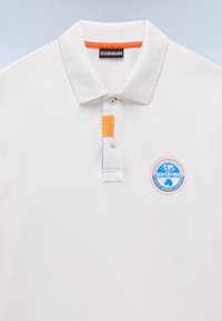 Valkoinen polo-paita, jossa on tekstuuri, oranssi kauluksen reuna, kaksi nappia ja pyöreä sininen logo-patch vasemmassa rinnassa.