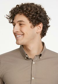 Cămașă button-up de culoare maronie deschisă, cu o textură moale, având un guler clasic și nasturi albi, purtată de un model cu păr creț.