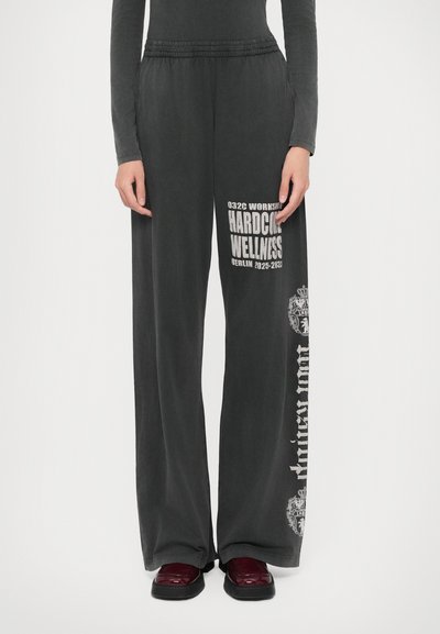 Pantaloni largi negri cu imprimeuri albe "Hardcore Wellness" și "Antikguom", purtați cu pantofi roșii închisi și un top negru cu mâneci lungi, vedere din față.