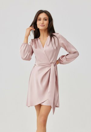 Kobieta o długich ciemnych włosach, ubrana w jasnoróżową satynową sukienkę kopertową z długimi rękawami, pozująca z jedną ręką przy twarzy, a drugą na biodrze.