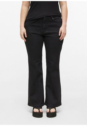 Osoba nosząca czarne jeansy bootcut i czarne sandały na platformie, z różowo pomalowanymi paznokciami u stóp, stojąca na jednolitym białym tle.