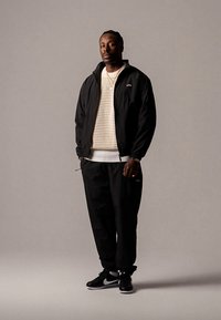 Črna jakna z zadrgo čez bež mrežast pulover, v kombinaciji s črnimi trenirkami in črnimi supergami. Opazni poudarki vključujejo belo plast spodaj.