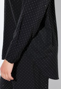 Blusa nera con pois argentati, maniche lunghe, polsini abbottonati e orlo curvo. Visibile una texture liscia e un tessuto leggero.