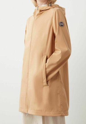 Mujer con chaqueta color beige con capucha y cremallera, con bolsillos laterales y un parche con el logo de Colmar en la manga izquierda, sobre un fondo liso.