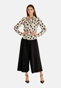 Langarmbluse in hellem Beige mit schwarzem Pinselstrich-Muster, kombiniert mit weit geschnittenen schwarzen Hosen und schwarzen spitzen High Heels.