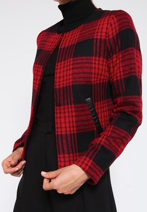 Giacca a quadri rossa e nera con maniche lunghe sopra un dolcevita nero, indossata da una persona che aggiusta l'orlo della giacca.