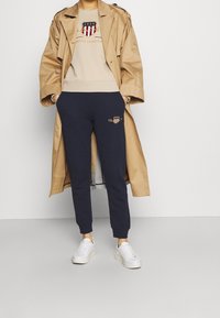 Trench-coat beige sur un sweat-shirt beige avec logo, associé à un pantalon de survêtement bleu marine avec un emblème brodé et des baskets blanches.