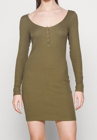 Kvinna i en olivgrön, långärmad stickad klänning med rundad halsringning och fyra små knappar framtill.