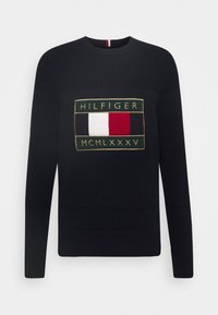 Granatowy sweter dzianinowy z wyraźnym haftowanym logo Hilfiger w prostokątnym wzorze z czerwonymi, białymi i niebieskimi akcentami.