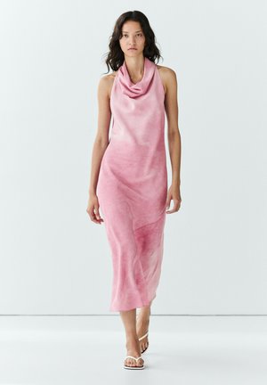 Kvinde gående i en ærmeløs pink midi-kjole med rullekrave og hvide sandaler med åben tå mod en ensfarvet hvid baggrund.