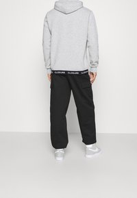 Grijze hoodie met een zicht op de achterkant, gecombineerd met zwarte cargobroeken met een logo op de tailleband. Schoenen zijn grijs met witte accenten.