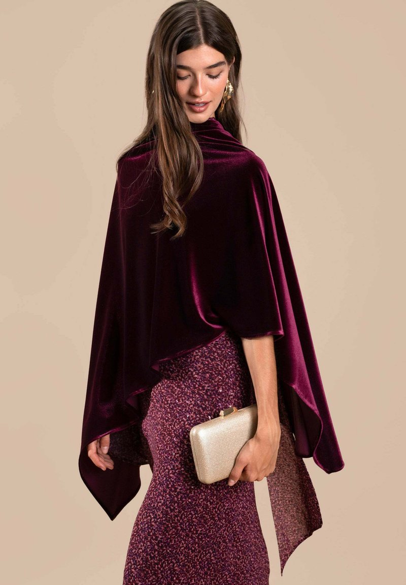 Capa de terciopelo burdeos con un diseño fluido y dobladillo puntiagudo, combinada con una falda púrpura texturizada y un pequeño bolso de mano dorado.