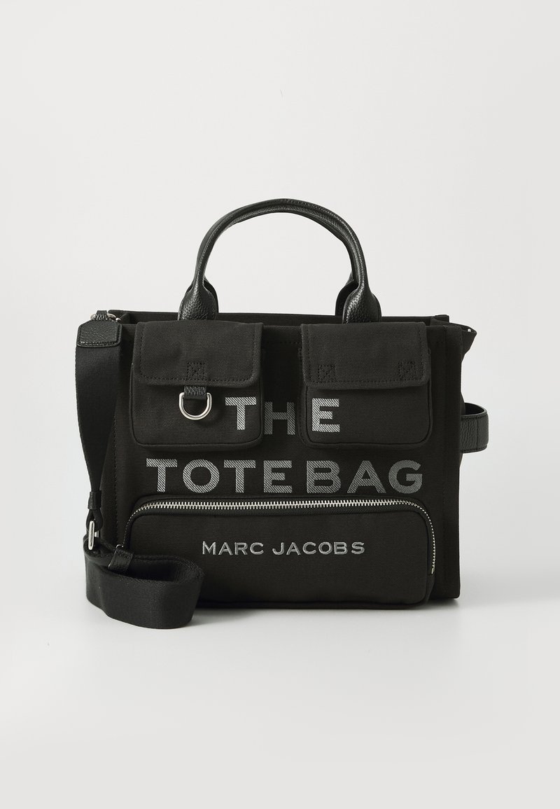 Bolsa tote preta da Marc Jacobs com dois bolsos frontais com aba, um compartimento com fecho e "THE TOTE BAG" impresso em texto branco.