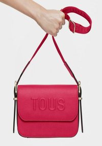 Bolso de mano de cuero rojo con el logo "TOUS" en relieve, forma rectangular, correa ajustable, herrajes en tono dorado y acentos en negro contrastantes.