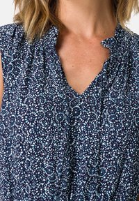 Blouse sans manches bleu marine avec un motif floral en bleu clair et blanc. Présente un col plissé et une texture de tissu douce.