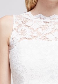 Vit klänning med en spetsöverdel som har blommönster, hög krage och är ärmlös. Slät textur under spetsen.