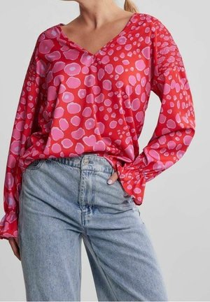 Blusa rossa con motivi circolari rosa, scollatura a V e maniche arricciate, abbinata a jeans a vita alta di blu chiaro.