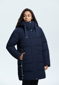 Marinblå pufferjacka med hög krage, justerbara dragsko, sidofickor med dragkedja och en boxig siluett. Slät, quiltad textur.