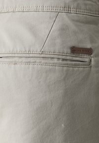 Pantaloni bej din bumbac, cu un buzunar pe spate prevăzut cu fermoar, accentuați de o mică etichetă din piele maro, cu inscripția "JACK & JONES" embosată.