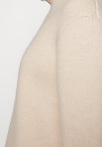 Beige Strickpullover mit hohem Kragen, der eine weiche Textur und gleichmäßige, feine Maschen aufweist, und das Design der Schulter- und Seitenansicht zeigt.